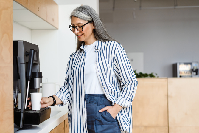 Happy white-haired mature woman smiling while using coffee maker at the office